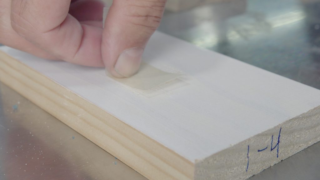 Applying tape to a white coated sample, preparing for the final step of the cross hatch adhesion test